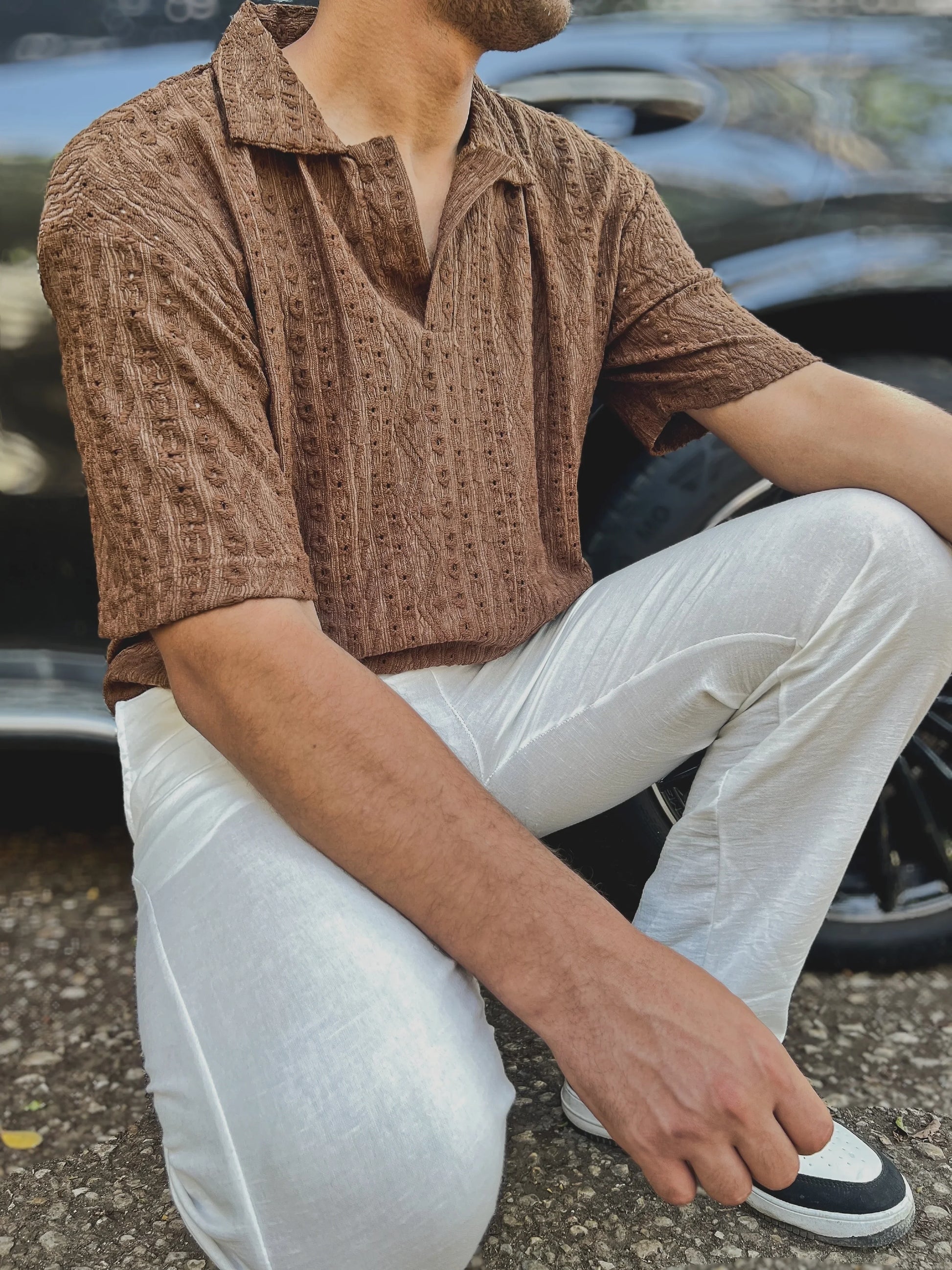 Person wearing a brown patterned shirt and white pants sitting on the ground.